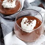 Horizontal image of two glass cups filled with a thick, dark brown dessert topped with whipped cream, on a towel next to metal spoons.