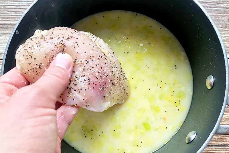 Horizontal image of a hand putting a piece of raw seasoned meat into a pot filled with a yellow liquid.