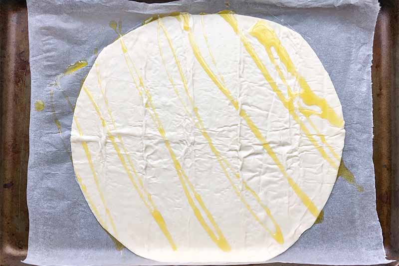 Horizontal image of an unbaked round flat dough drizzled with olive oil on a baking sheet.