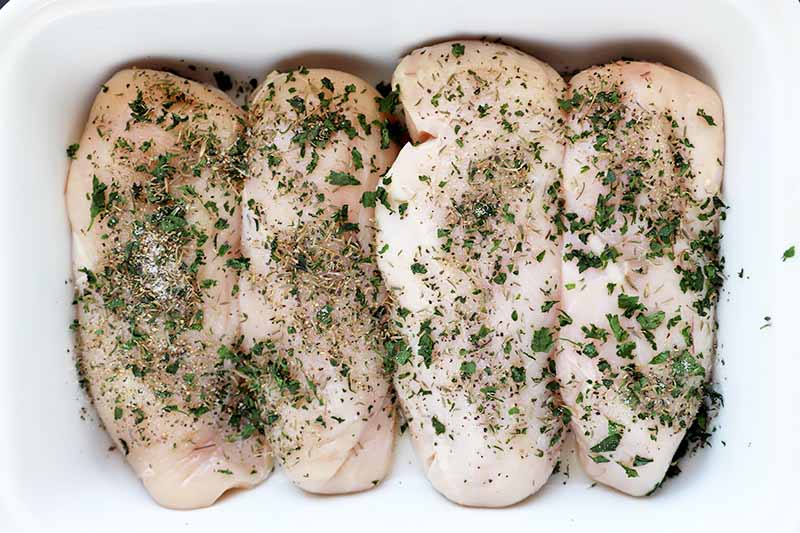 Horizontal image of four raw poultry breasts covered in seasonings in a white dish.