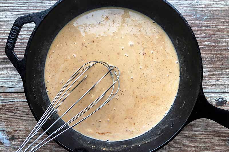 Horizontal image of a very light red creamy liquid stirred with a metal whisk in a pan.