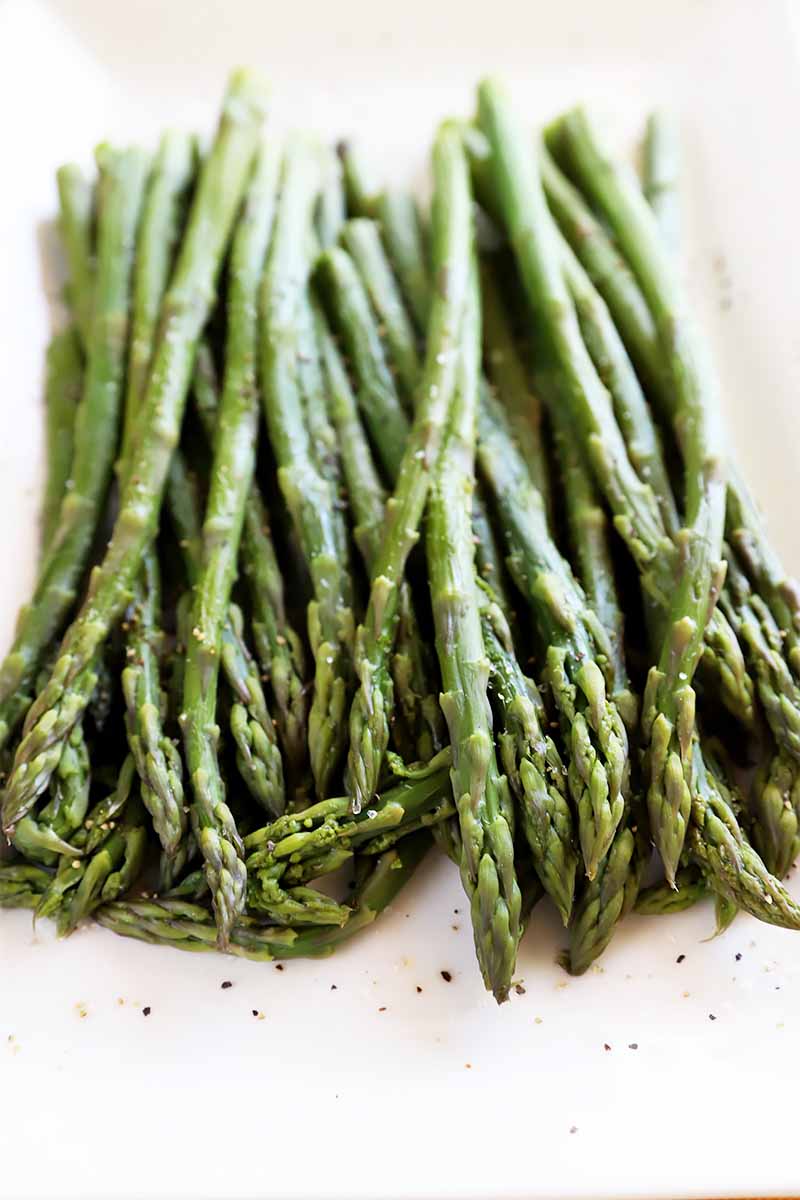 Vertical close-up image of cooked green vegetable spears on a white plate with cracked black pepper.