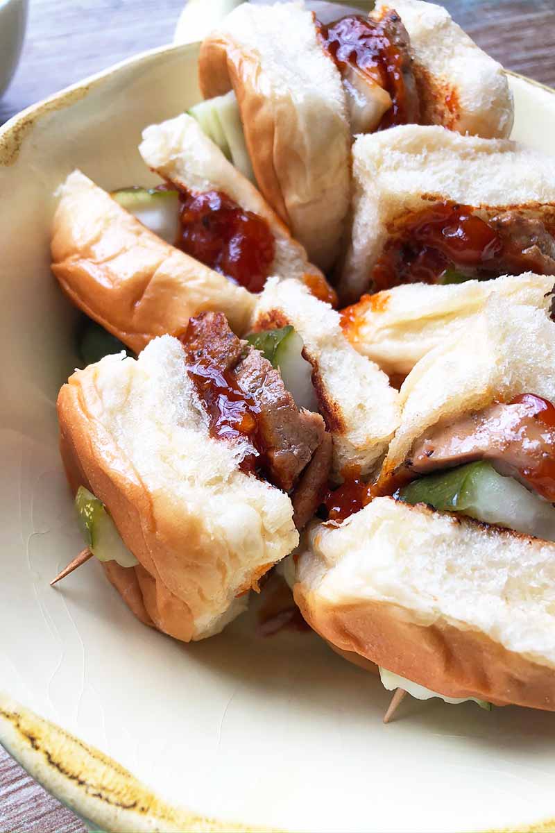 Vertical close-up image of a white platter with small meat sandwiches with pickles and a thick red sauce.