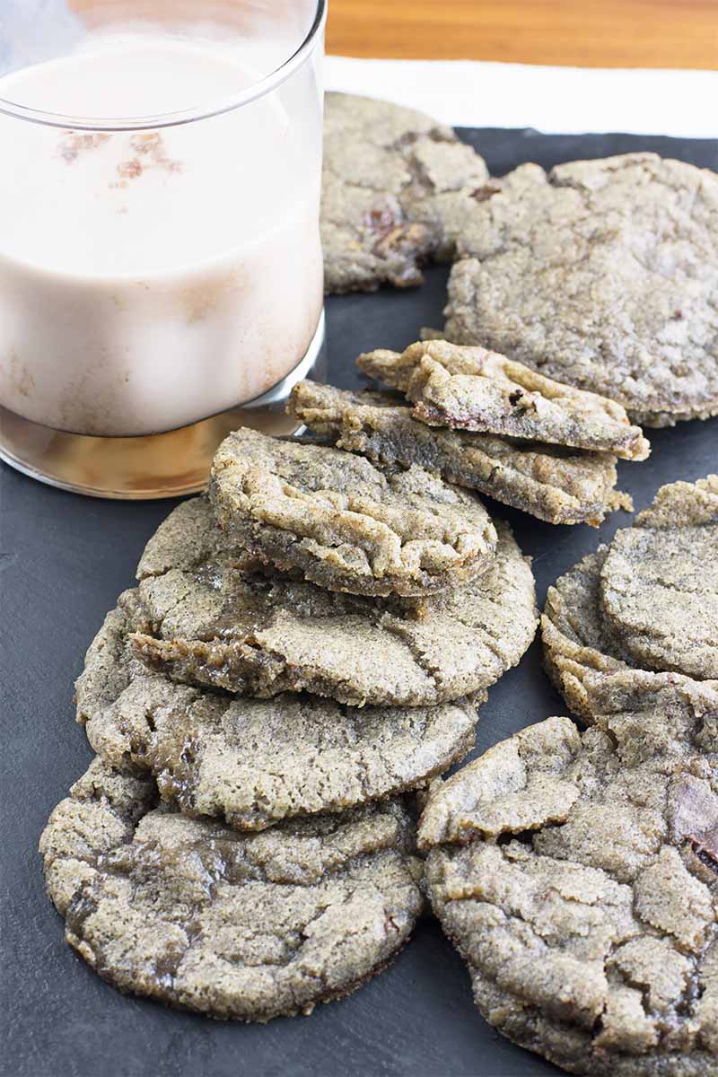 Vertical image of a mound of cookies on a slate next to a glass of milk.
