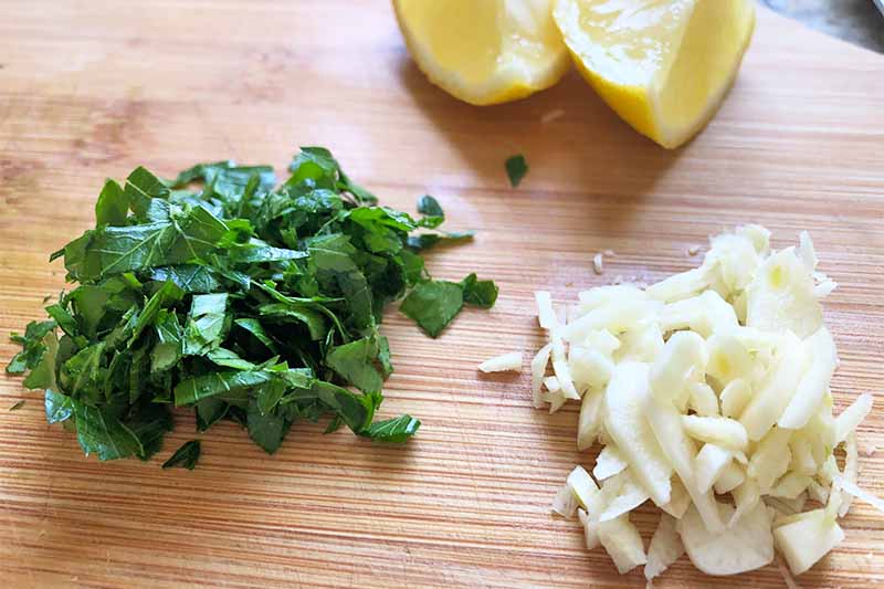 Horizontal image of chopped herbs, garlic, and wedges of citrus on a wooden board.