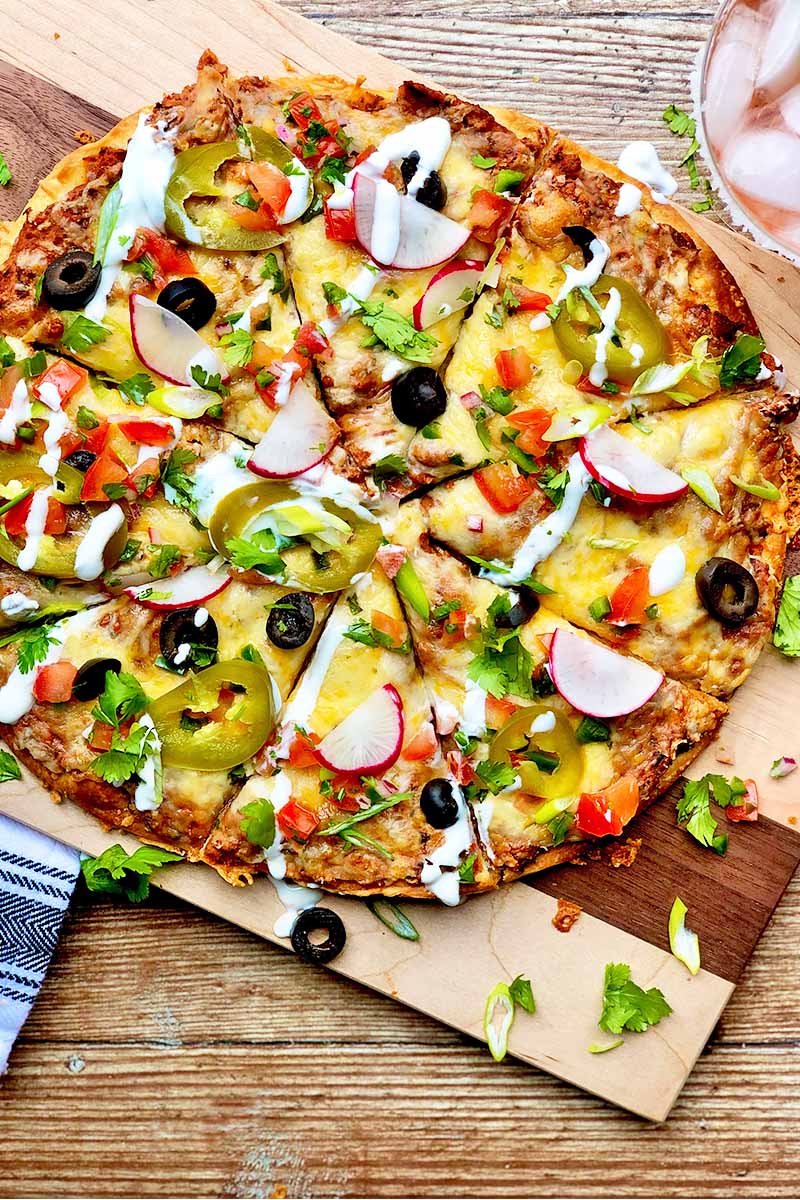 Vertical top-down image of a sliced round cheesy flatbread garnished with mayo and assorted colorful toppings on a wooden cutting board.