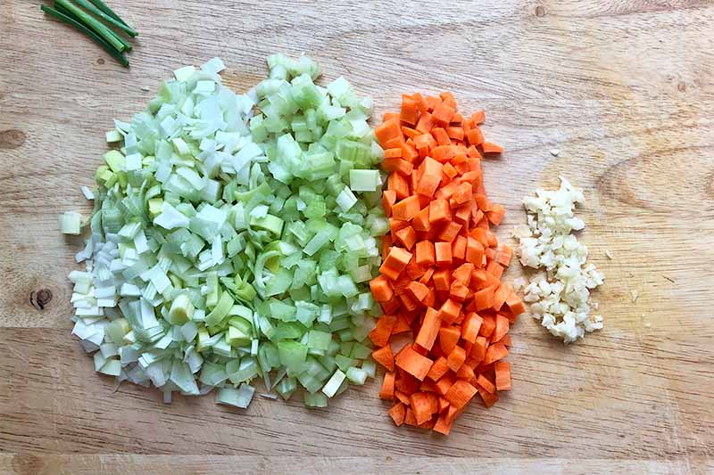 Horizontal image of chopped onions, celery, carrots, and garlic on a wooden cutting board.