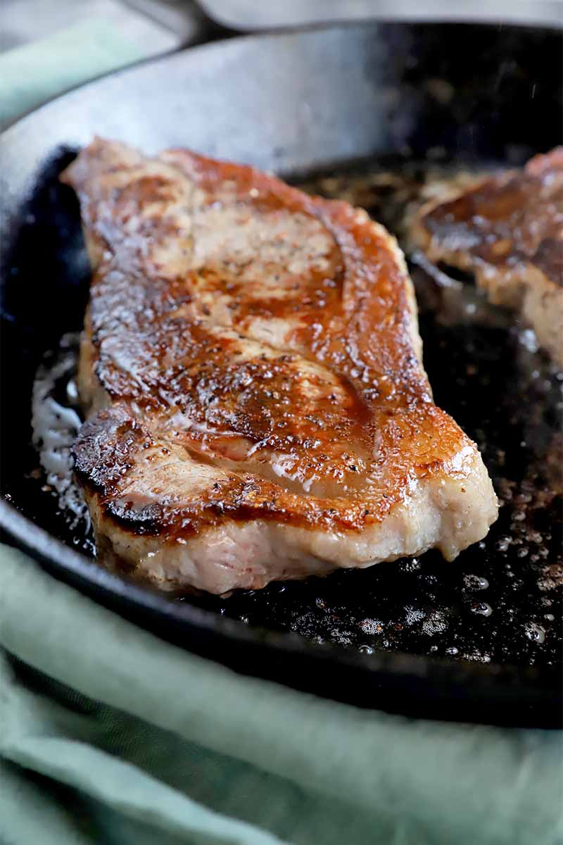 Vertical image of a thick cut of meat browned in a cast iron skillet with melted butter and salt and pepper.