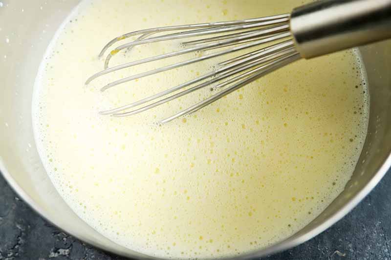 Horizontal image of a boiled milk mixture stirred by a metal whisk in a metal bowl.