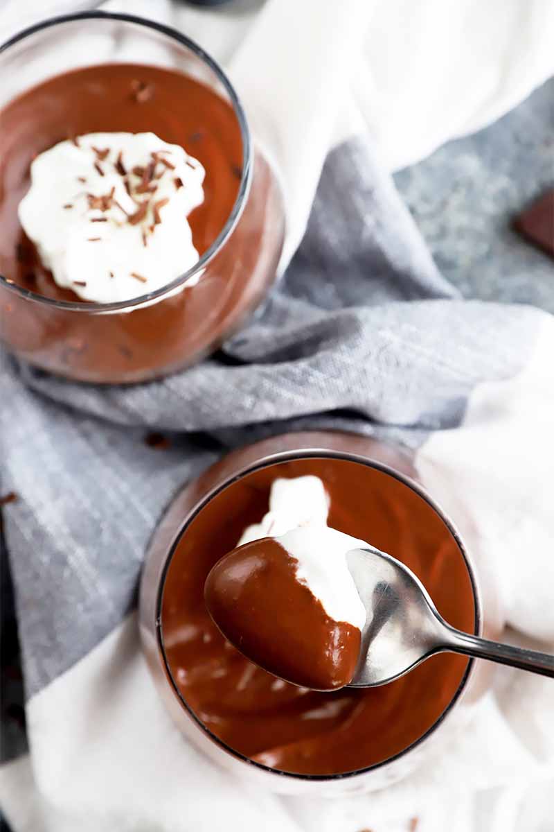 Vertical image of metal spoon holding a spoonful of a chocolate pudding with whipped cream next to the same dessert on a towel.