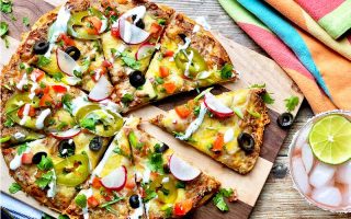 Horizontal image of slices of a round flatbread covered in assorted colorful fresh toppings and a drizzle of mayonnaise on a wooden cutting board next to a cocktail and a colorful towel.