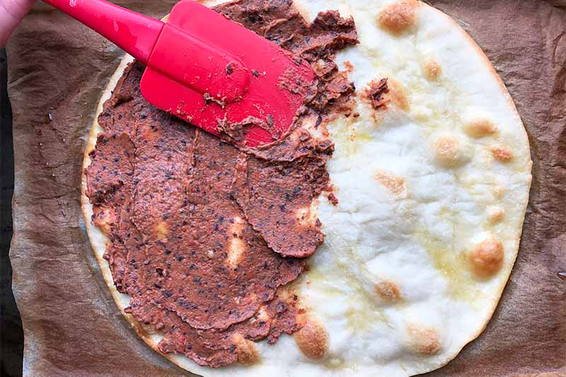 Horizontal image of a red spatula spreading a dark brown mixture on top of a slightly baked round flatbread.