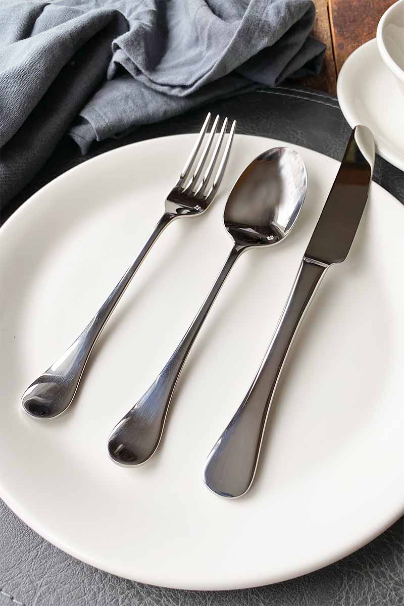 Vertical image of a metal fork, spoon, and knife on a white plate next to gray napkins.
