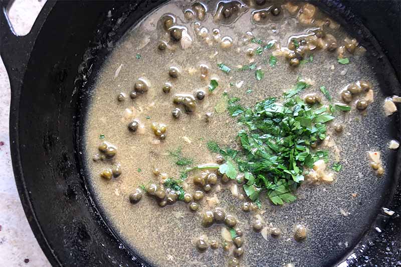 Horizontal image of cooking a sauce with capers and a mound of fresh chopped herbs.
