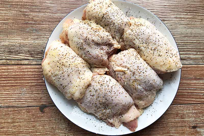 Horizontal image of a white plate with 6 pieces of raw, skin-on poultry with seasoning.