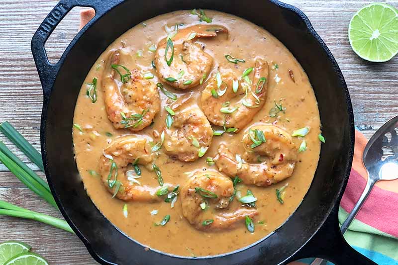 Horizontal image of a cast iron skillet filled with cooked shrimp in a creamy sauce garnished with sliced spring onions.