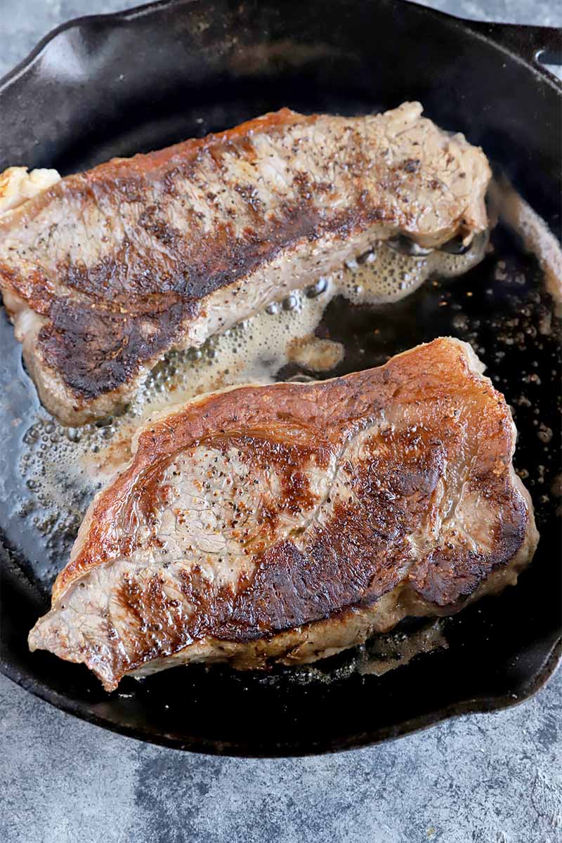 Vertical image of two thick cuts of beef seasoned and browned in a skillet with melted butter.