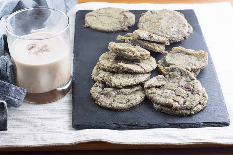 Horizontal image of partially stacked cookies on a slate platter on a white towel next to a glass of milk.