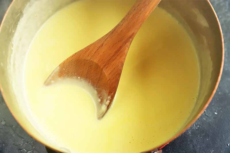 Horizontal image of a thick milk mixture stirred by a wooden spoon in a metal bowl.
