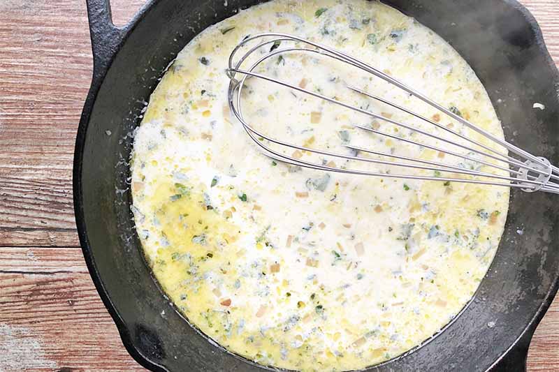 Horizontal image of whisking a thick, white liquid with chopped herbs in a cast iron skillet.