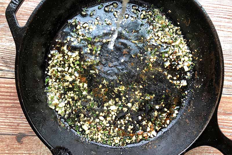 Horizontal image of pouring liquid in a pan with cooked aromatics.