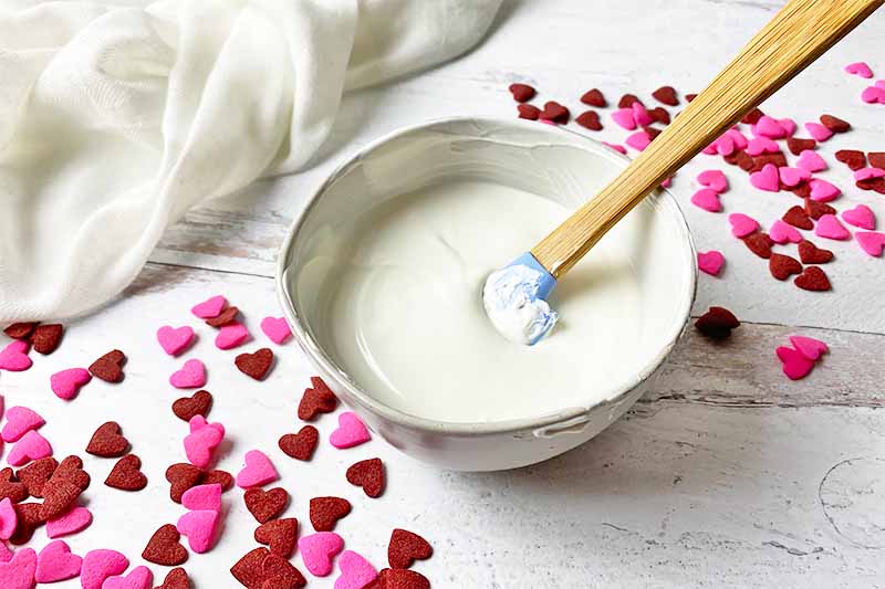 Horizontal image of melted white candies in a white bowl stirred by a spatula with Valentine's Day themed chocolate hearts on the surface.
