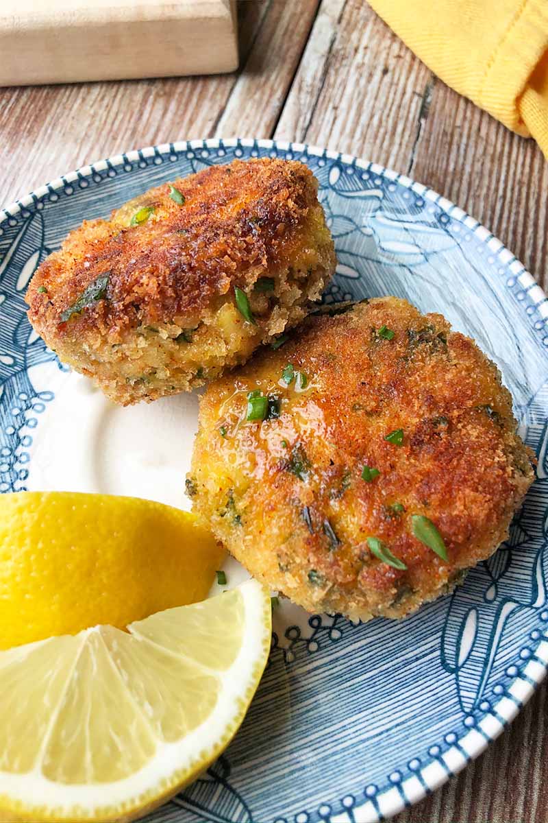Vertical image of two breaded and browned patties with fresh herbs on a plate with lemon wedges on a wooden table.
