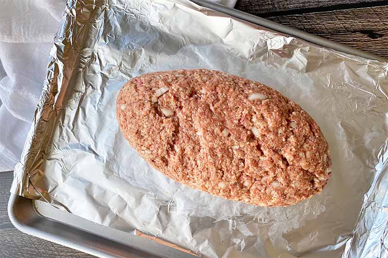 Horizontal image of a raw, oblong mound of ground beef on a baking pan lined with aluminum foil.