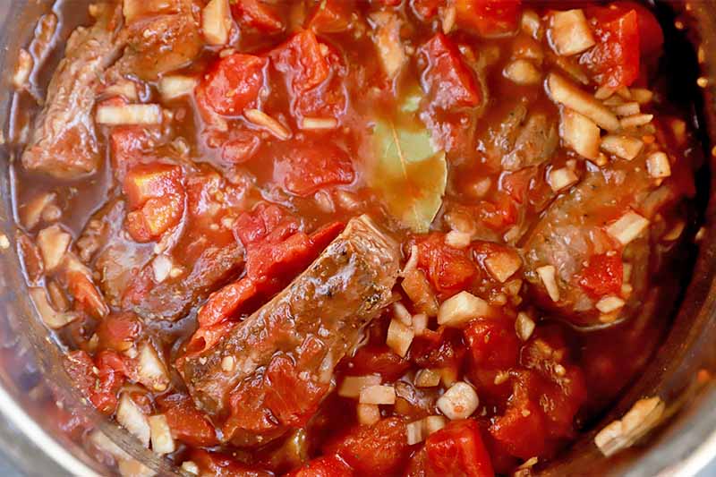 Horizontal image of tomato sauce, onions, a bay leaf, and chunks of meat in a large bowl.