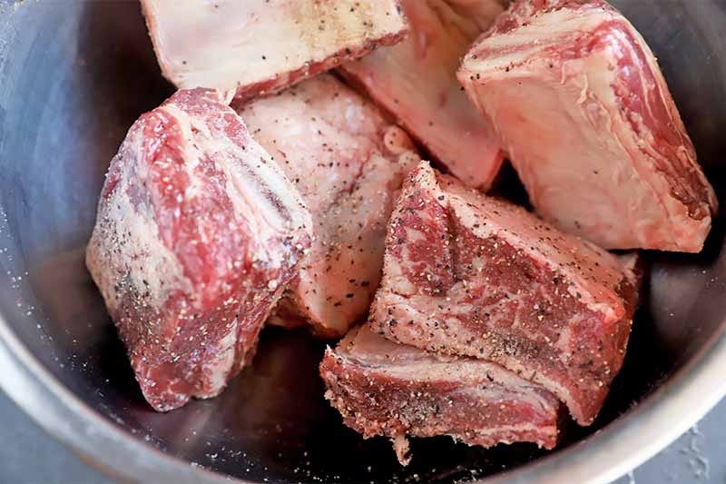Horizontal image of raw chunks of meat seasoned with salt and pepper in a bowl.