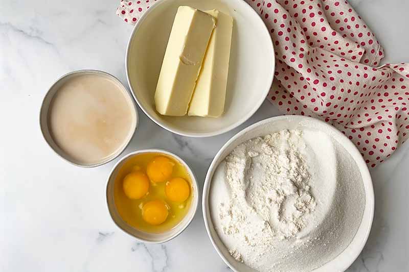 Horizontal image of bowls of milk, eggs, butter, and dry ingredients next to a towel with polka dots.