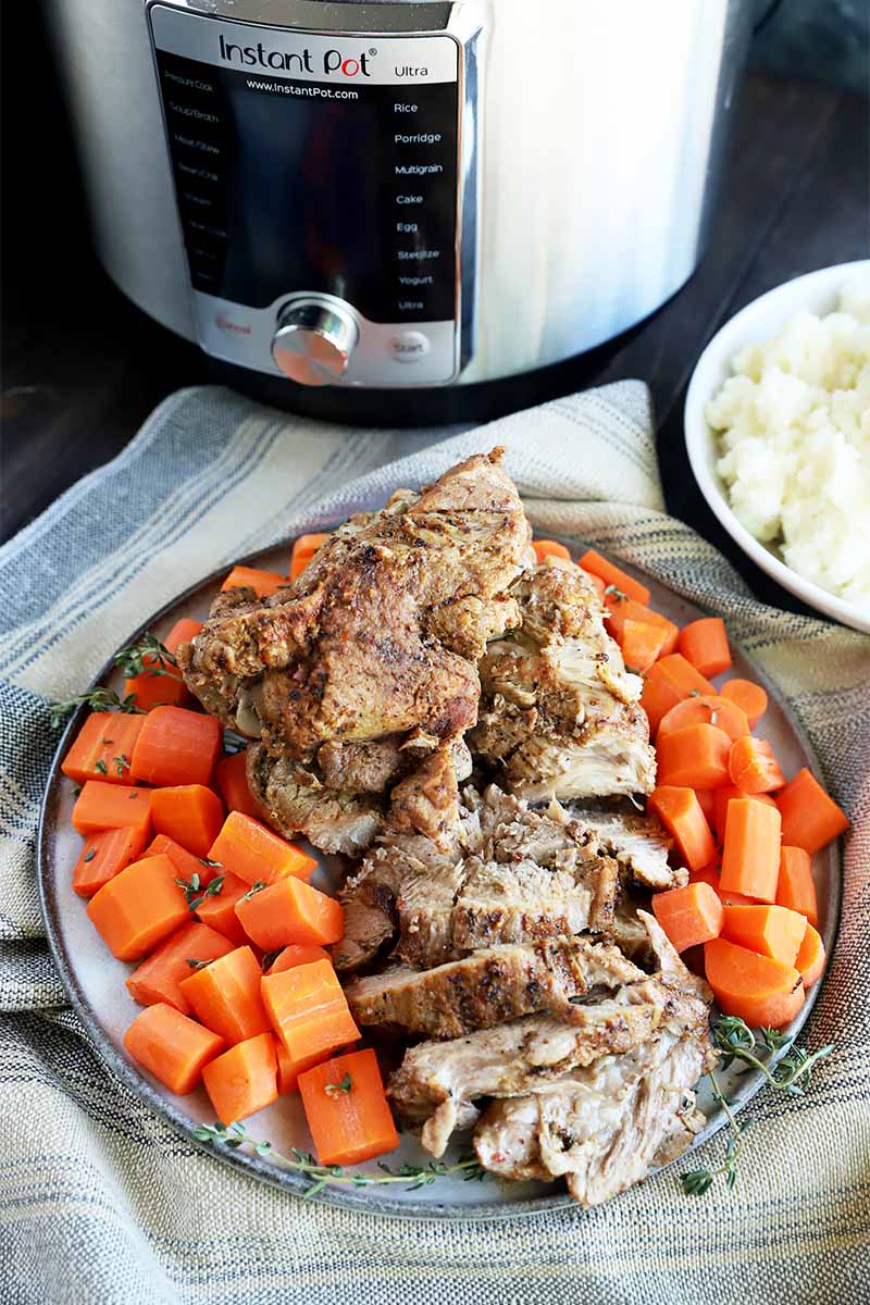 Vertical image of a large plate full of chopped cooked carrots and sliced cooked meat on top of a tan towel in front of a kitchen appliance.
