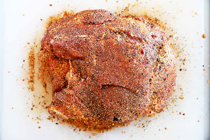 Horizontal image of a large piece of raw meat rubbed with a dry spice mixture on a cutting board.