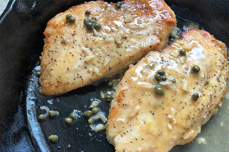 Horizontal image of two seared poultry breasts in a sauce with capers and minced garlic in a cast iron skillet.