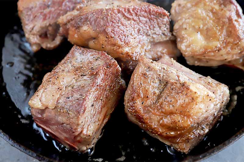 Horizontal image of seared large chunks of seasoned meat cooking in an oiled skillet.