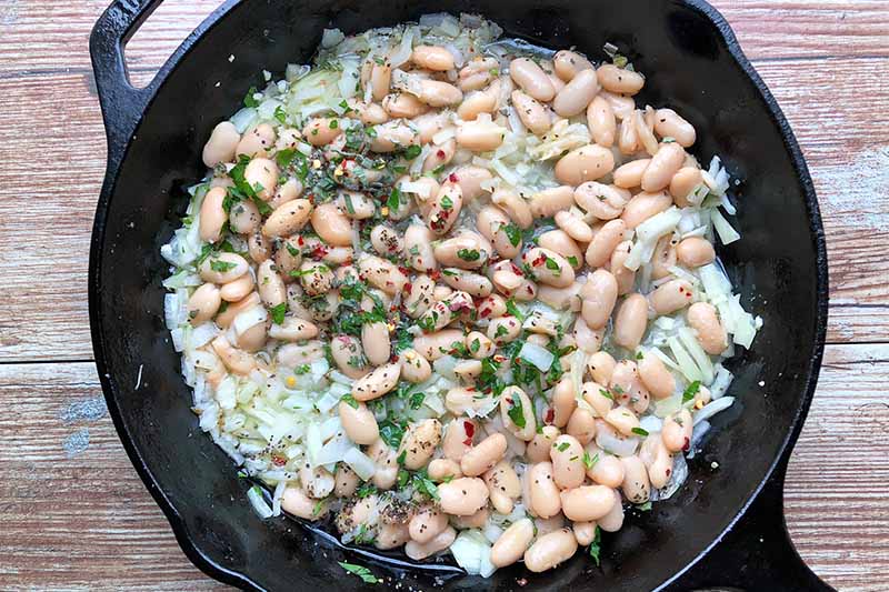 Horizontal image of cooking white beans, herbs, and chopped onions in a cast iron skillet.