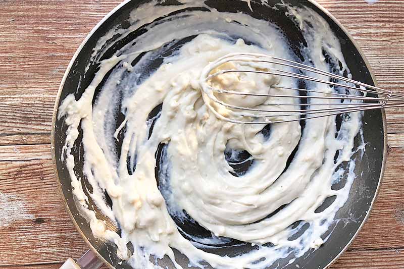 Horizontal image of a very thick white sauce stirred with a whisk in a pan.