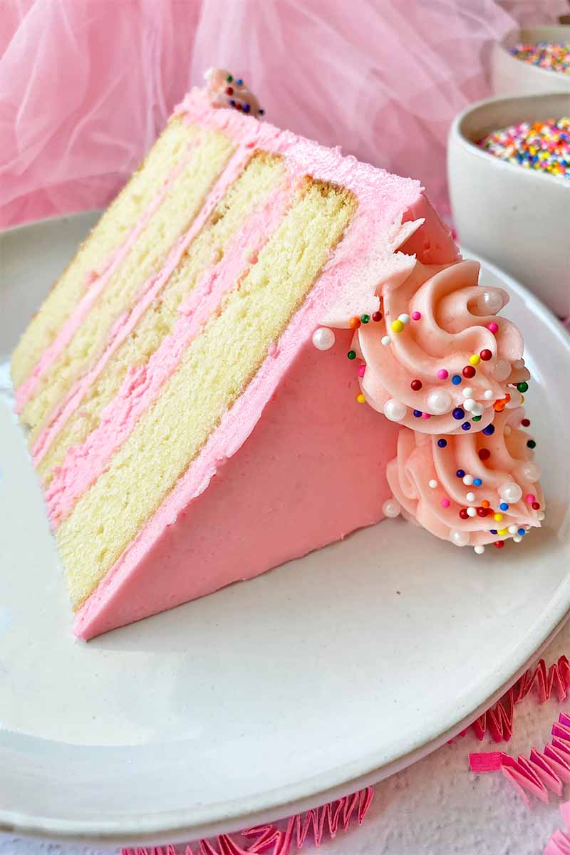 Vertical image of a slice of vanilla dessert with colorful frosting and sprinkles on a white plate.