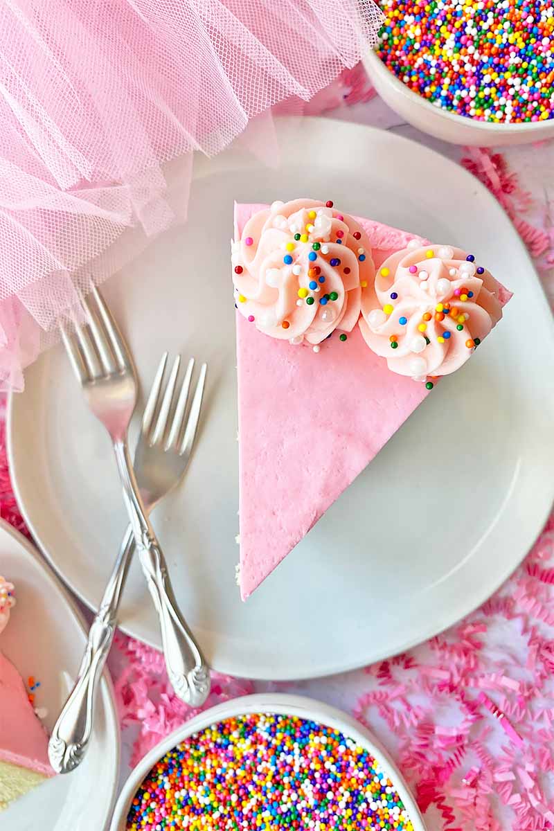Vertical top-down image of a triangular slice of a pink dessert on a white plate next to bowls of sprinkles, forks, and confetti.