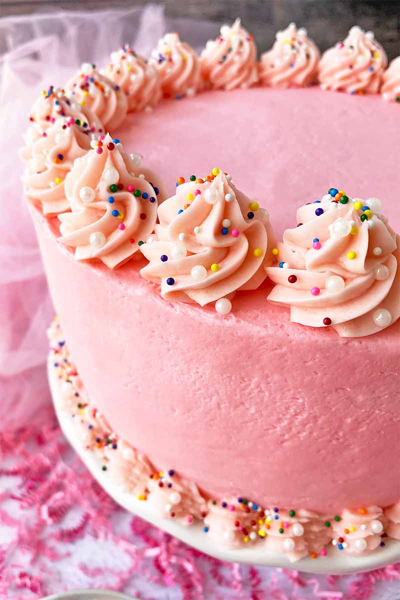 Vertical close-up image of a decorated dessert with light red frosting and sprinkles on a white stand surrounded by confetti.