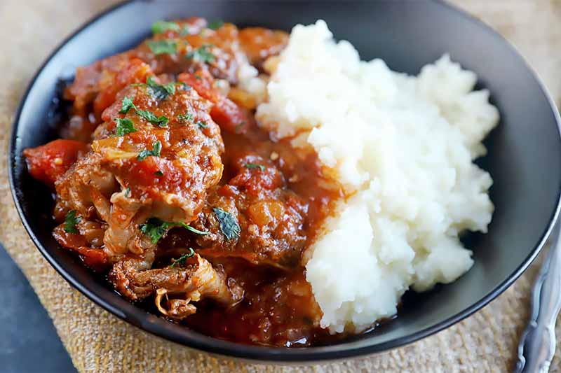 Horizontal image of a black bowl filled with chunks of cooked meat in a tomato sauce next to mashed potatoes, all on top of a brown mat.