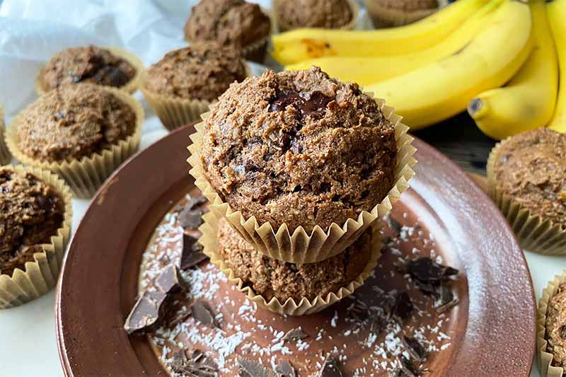 Horizontal image of a stack of muffins on a brown plate in front of more baked goods and bananas.