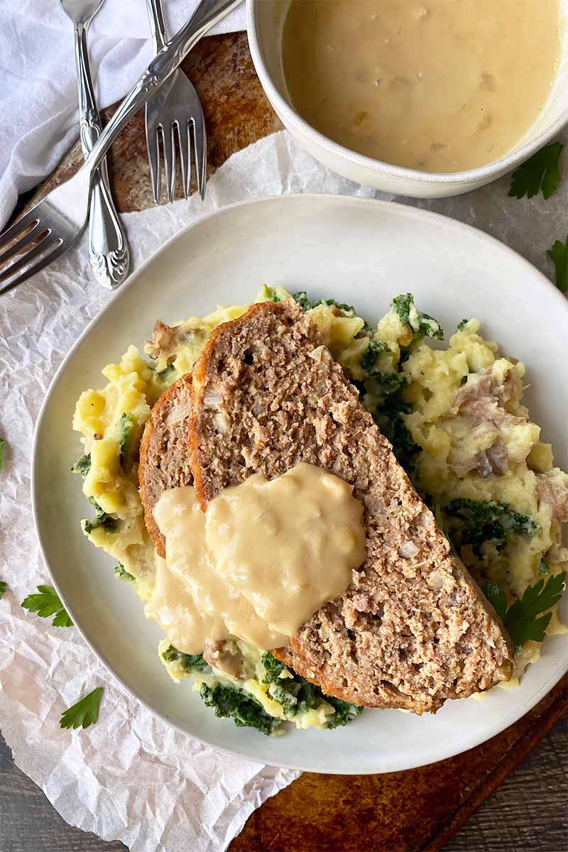 Vertical top-down image of two slices of a ground beef dish on top of mashed kale potatoes served with gravy on a white plate.