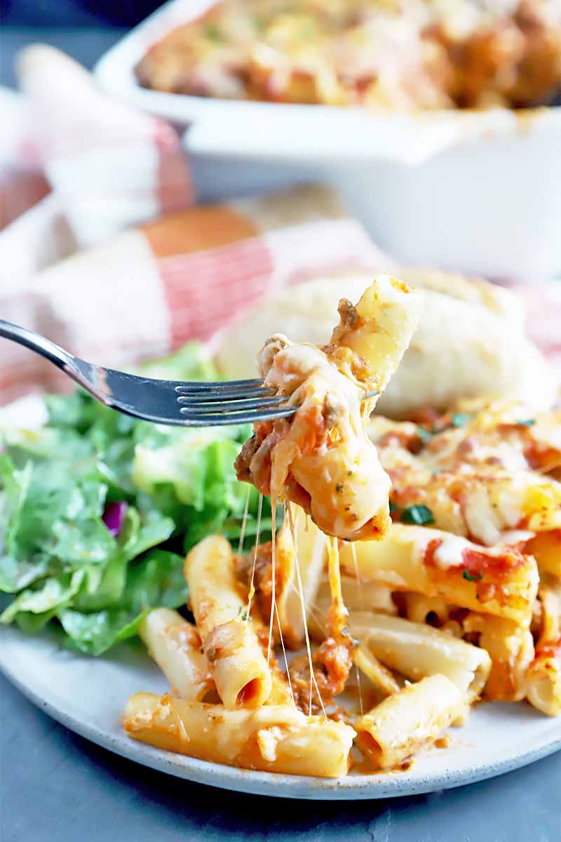 Vertical image of a fork holding a very cheesy piece of a baked casserole dish on a plate with salad on a red and white checkered towel.