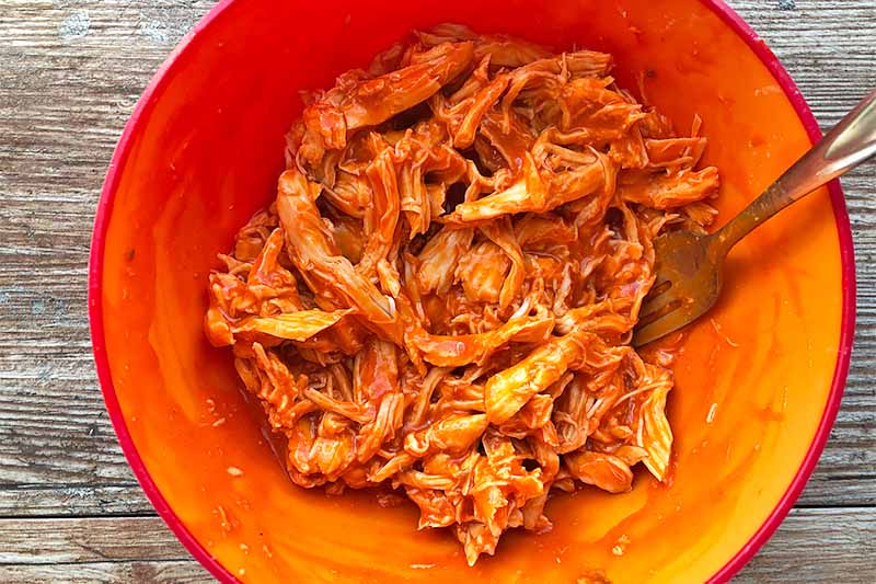 Horizontal image of shredded meat in a red sauce in a red bowl.