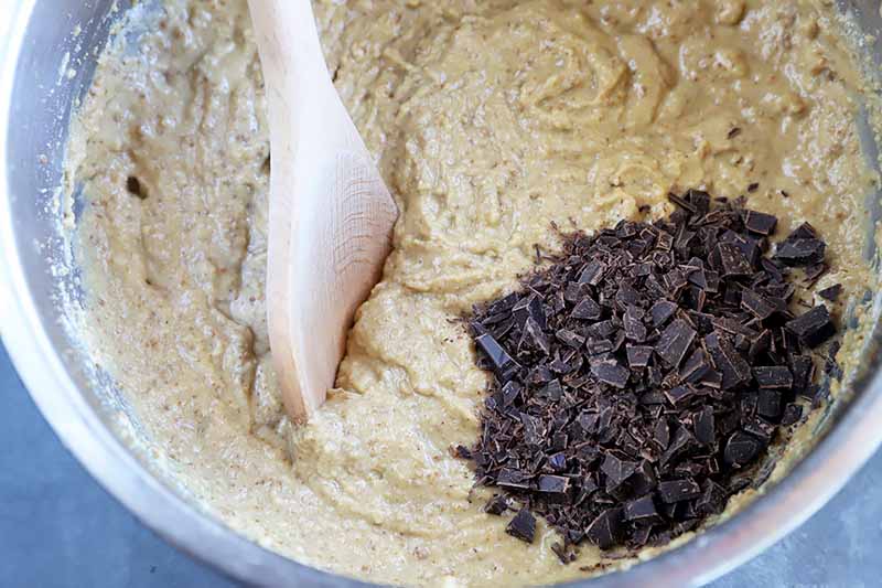 Horizontal image of stirring in a large clump of chocolate chunks into a light yellow batter with a wooden spoon.