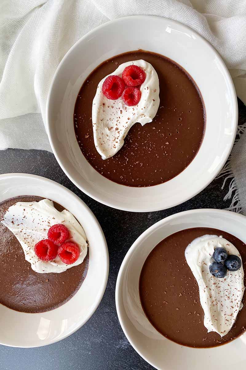 Vertical top-down image of three white bowls filled with chocolate pudding topped with whipped cream and fruit.