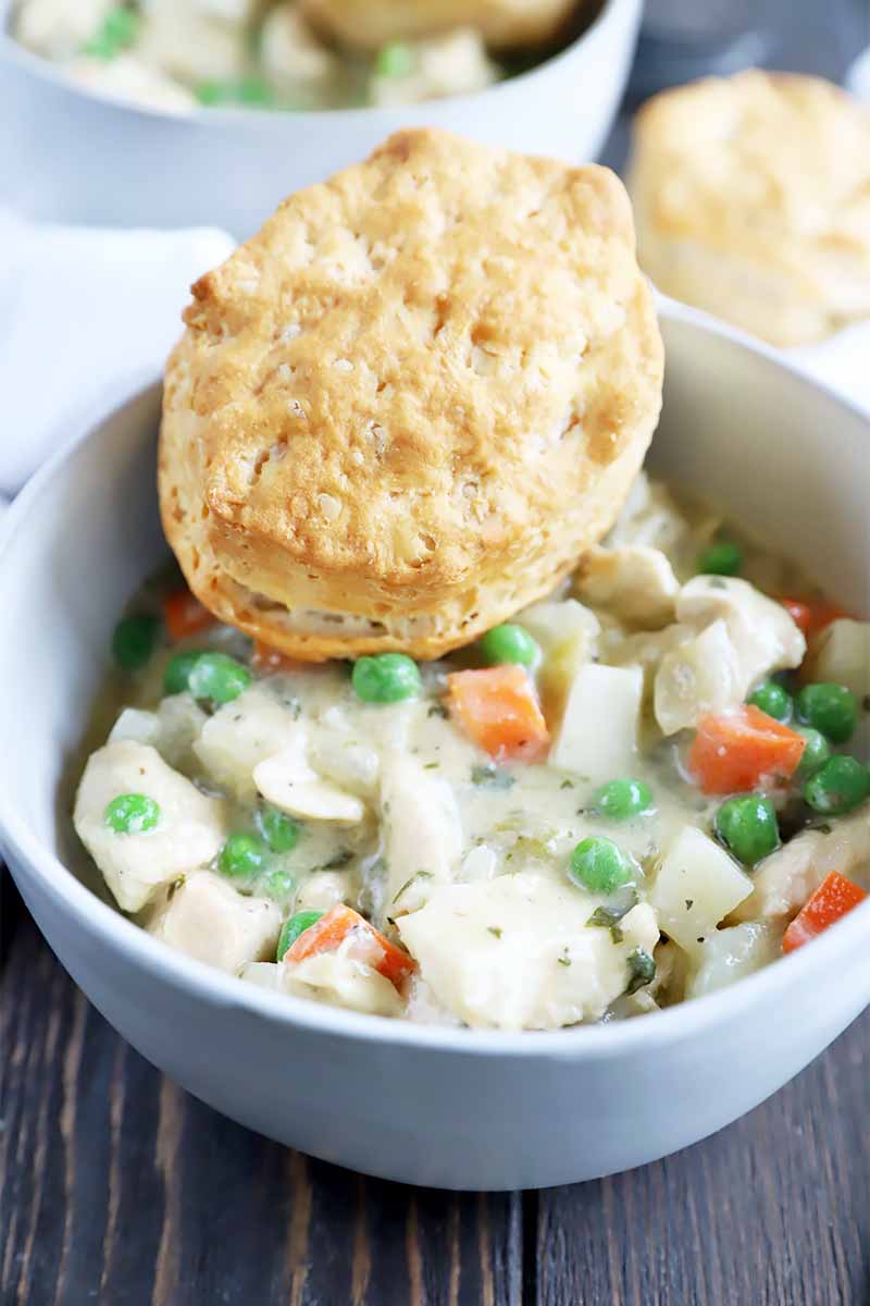 Vertical Image of a white bowl filled with a creamy mixture of vegetables and white meat with a biscuit on top.