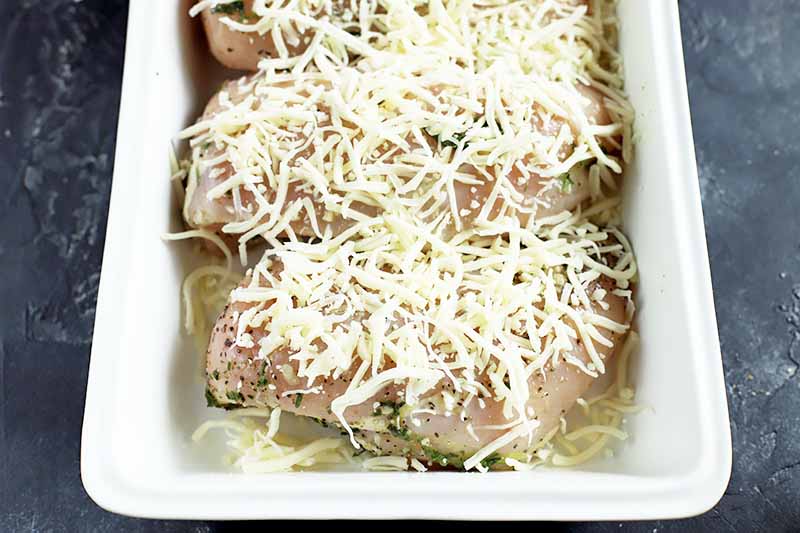 Horizontal image of uncooked poultry breasts covered in shredded white cheese and an oil/herb mixture.