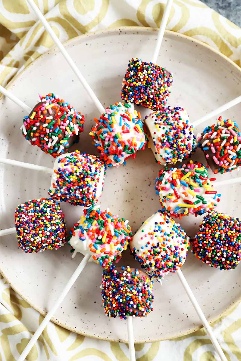 Vertical image of marshmallows on sticks covered in assorted sprinkles on a white plate.